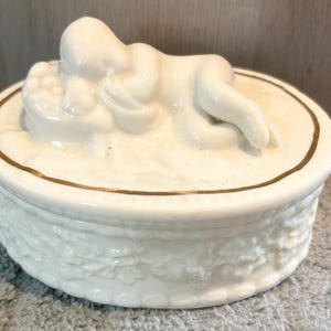 Puede incluir: Caja de baratijas de cerámica blanca con tapa que presenta una figura de bebé esculpida. La caja tiene un borde dorado y un diseño en relieve texturizado alrededor de la base. El bebé está acostado en la tapa.