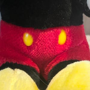 May include: A plush black and red Mickey Mouse toy with yellow buttons. The toy is sitting with its legs crossed.