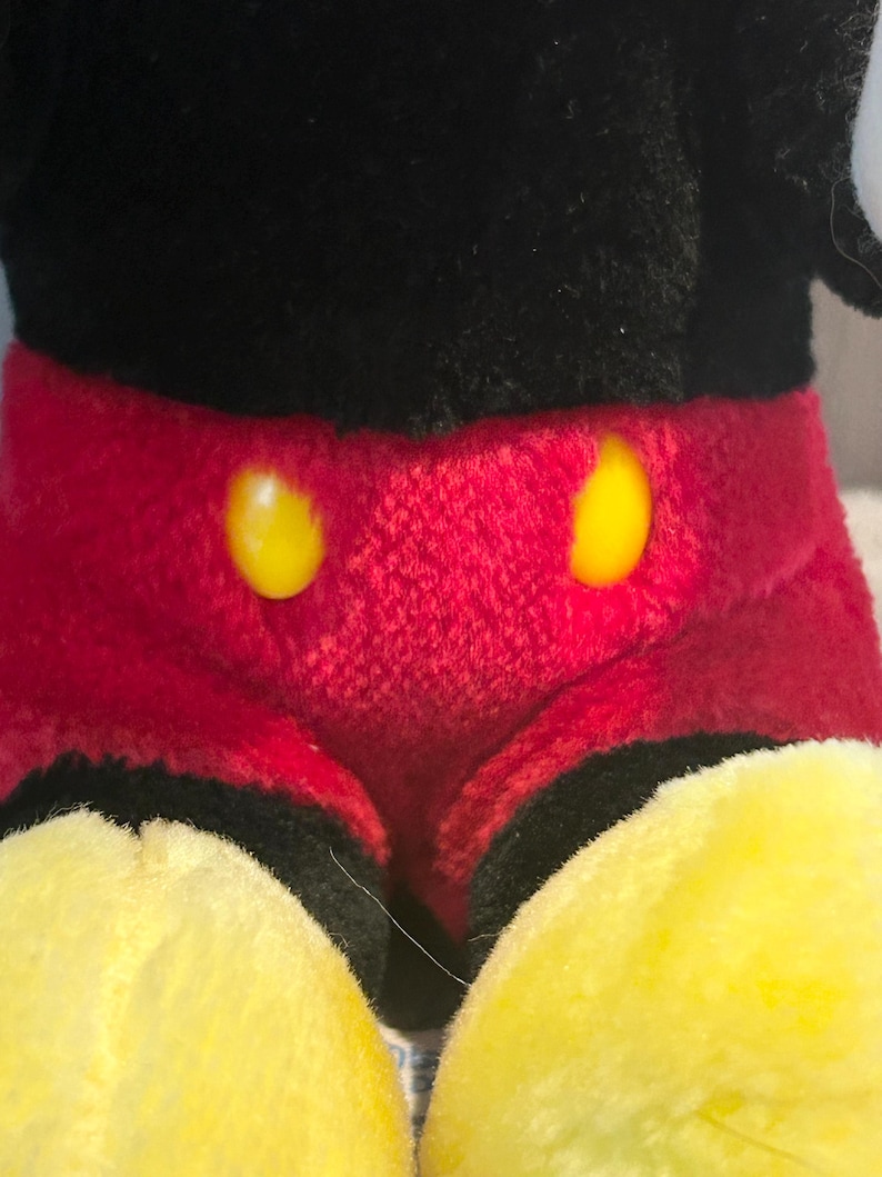 May include: A plush black and red Mickey Mouse toy with yellow buttons. The toy is sitting with its legs crossed.