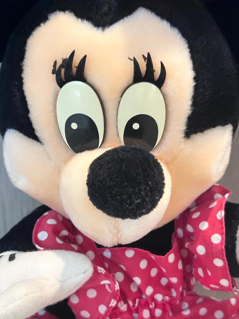 May include: A plush toy of a cartoon mouse wearing a pink dress with white polka dots. The mouse has large black eyes and a black nose.
