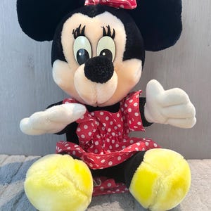 May include: A plush Minnie Mouse toy wearing a red dress with white polka dots and a red bow. She has black ears, yellow feet, and a black nose.