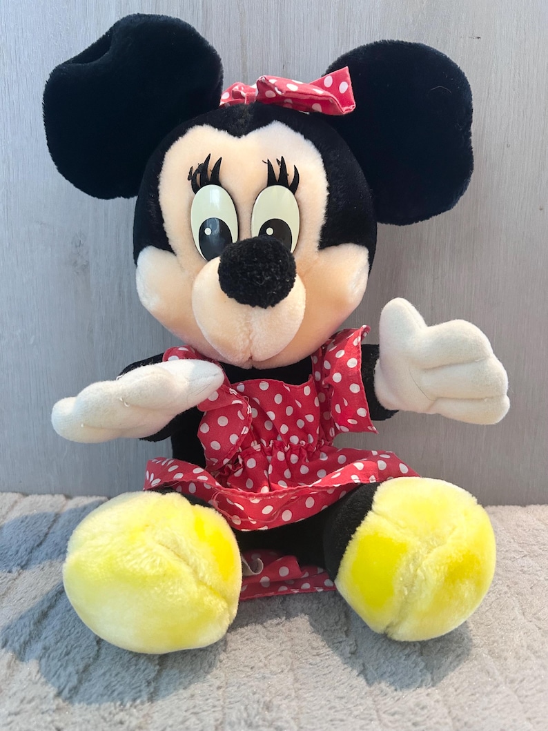 May include: A plush Minnie Mouse toy wearing a red dress with white polka dots and a red bow. She has black ears, yellow feet, and a black nose.