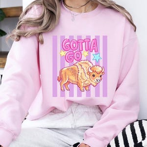 May include: A light pink sweatshirt with the phrase "Gotta Go" in purple and yellow text, along with a cartoon bison illustration. The bison is tan and brown, and the background has vertical purple stripes.