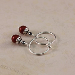 May include: A pair of silver hoop earrings with dangling red beads. Each earring features a dark red, round bead, a silver accent with star and crescent moon designs, and a small black bead. The hoops are silver and have a simple clasp.