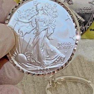 Może przedstawiać: Srebrny wisiorek w kształcie monety ze szczegółowym przedstawieniem Lady Liberty, napisami "IN GOD WE TRUST" i rokiem 2026. Moneta jest przymocowana do srebrnego breloka. Moneta ma teksturowaną, żebrowaną krawędź.