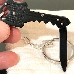 May include: A black key-shaped multi-tool with the brand name "SOG" visible. The tool features a folding knife blade and is attached to a silver keyring with a chain. The key-shaped tool is held open, displaying the knife blade.