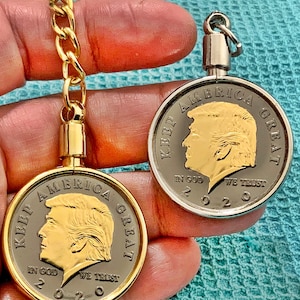 Pode incluir: Dois chaveiros dourados com uma moeda preta e dourada que apresenta o perfil de um homem com o texto "KEEP AMERICA GREAT" e "IN GOD WE TRUST 2020".