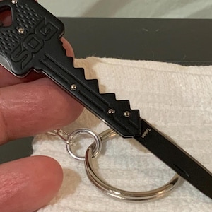Op de afbeelding: Zwart SOG vouwmes in sleutelvorm met een zilveren sleutelring. Het mesblad is uitgeschoven en toont een scherpe rand. De sleutelvormige handgreep heeft het SOG-logo en een gekartelde rand. Het mes is bevestigd aan een zilveren sleutelring.