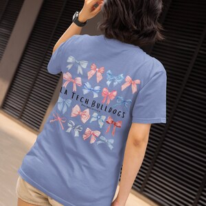 Pode incluir: Uma camiseta azul claro com um gr&aacute;fico de la&ccedil;os rosa e azul e o texto "LA Tech Bulldogs" nas costas.