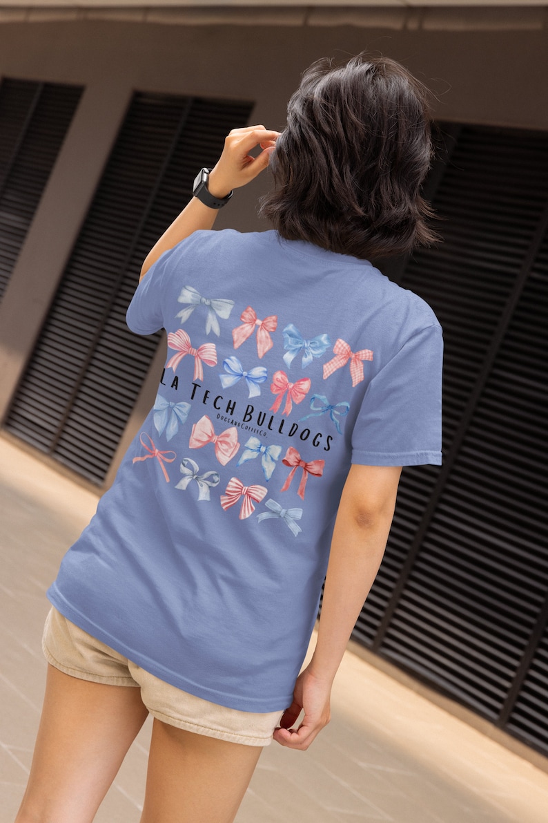 Pode incluir: Uma camiseta azul claro com um gr&aacute;fico de la&ccedil;os rosa e azul e o texto "LA Tech Bulldogs" nas costas.