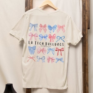 Pode incluir: Uma camiseta bege com um gr&aacute;fico de la&ccedil;os azuis e rosa e o texto "LA Tech Bulldogs" com as palavras "Dogs & Coffee Co." abaixo.