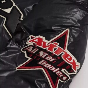 May include: Close-up of a black jacket with a glossy finish. The jacket features a large star-shaped patch with the text "Avirex All Star Dealers" in red and white. The jacket has a quilted design.
