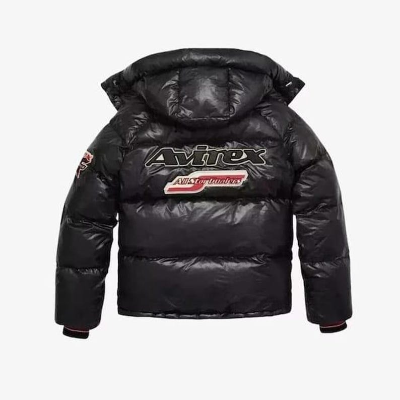 May include: Black puffer jacket with a hood. The jacket features the brand name "Avirex" in white and red lettering on the back, along with a patch. The jacket has a glossy finish and a quilted design.