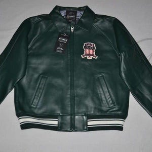 May include: Dark green leather jacket with a zip-up front, collared neckline, and a patch on the chest. The jacket has two front pockets and white stripes on the cuffs and hem. The jacket has the word "AVIREX" on the patch.