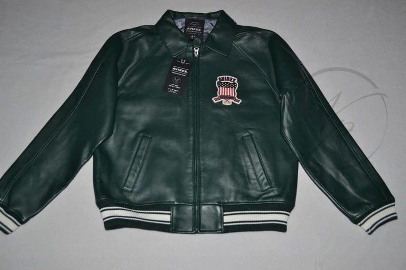 May include: Dark green leather jacket with a zip-up front, collared neckline, and a patch on the chest. The jacket has two front pockets and white stripes on the cuffs and hem. The jacket has the word "AVIREX" on the patch.