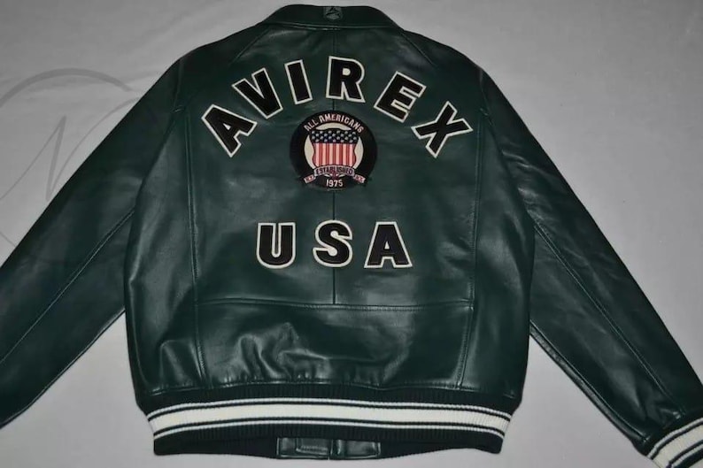 May include: Dark green leather jacket with white lettering spelling "AVIREX" and "USA" on the back. A circular patch with the American flag is centered between the words. The jacket has a white and black striped trim.