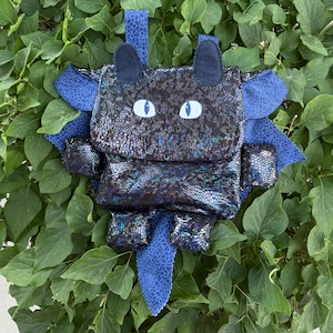 Op de afbeelding: Een handgemaakte rugzak met drakenmotief in een glinsterende zwart-blauwe stof. De rugzak heeft blauwe vleugels, oren en een staart, met witte en blauwe ovale ogen. De tas staat tegen een achtergrond van groene bladeren.