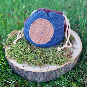 Könnte beinhalten: Ein blauer Filzbeutel mit Kordelzug, verziert mit einer braunen Tür, einer kleinen blauen Perle und einem beigen Kordelzug. Der Beutel liegt auf einem Bett aus grünem Moos auf einer Holzscheibe, mit einem grasbewachsenen Hintergrund.