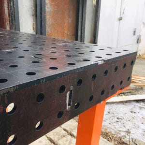 May include: A black metal welding table with numerous evenly spaced holes. The table is supported by bright orange legs. The surface is divided into a grid pattern. The table is designed for welding and metalworking projects.
