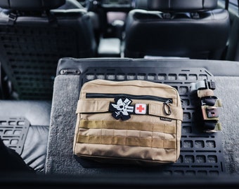 May include: A tan-colored medical bag with a black zipper and a black strap. The bag has a white patch with a skull and crossbones and the word "MEDIC" on it. There is also a red cross patch on the bag.
