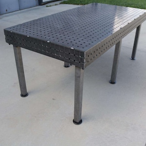 以下が含まれることがあります： 穴あき表面と4本の頑丈な脚を備えた長方形の金属製作業台。 テーブルは濃い灰色の金属製で、上部には等間隔に配置された穴のグリッドがあります。 脚には黒いゴム製の足が付いています。