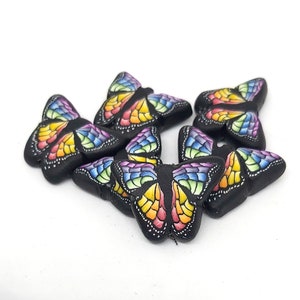May include: Five black butterfly-shaped polymer clay beads with rainbow painted wings. The beads are approximately 1 inch in size.