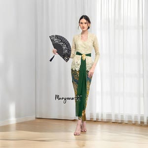 Peut inclure: Une femme portant un haut en dentelle blanche avec une jupe à motifs verts et dorés. Elle tient un éventail à main noir et porte une ceinture dorée à la taille.