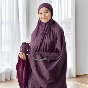 May include: A woman wearing a purple prayer shawl with a matching purple headscarf. She is holding a small purple pouch with a gold drawstring. The pouch has two small white beads hanging from it.