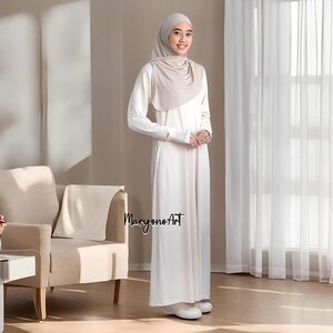May include: A woman wearing a cream-colored hijab and a matching long-sleeved dress. The dress has a simple design with a modest neckline and a flowing skirt. The text "Maryono Art" is visible on the dress.