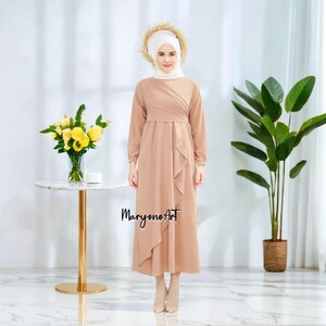 May include: A long-sleeved, light brown dress with a ruffled skirt and a white headscarf. The dress has a wrap-style design and a relaxed fit. The model is standing in front of a white wall with a potted plant and a table.
