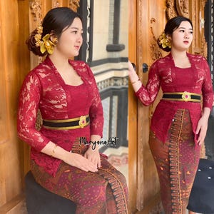 May include: A woman wearing a red lace top with a black belt and a red and gold patterned skirt. She is standing in front of a wooden door.