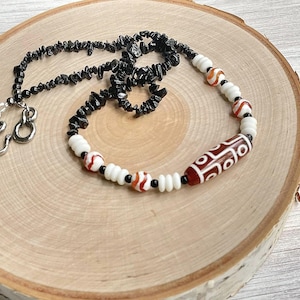 May include: A black and white beaded necklace with a red and white carnelian bead. The necklace is on a wooden surface.