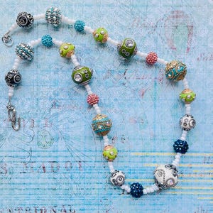 May include: A white beaded necklace with colorful, patterned beads. The beads are a mix of green, blue, pink, and black. The necklace has a silver clasp.