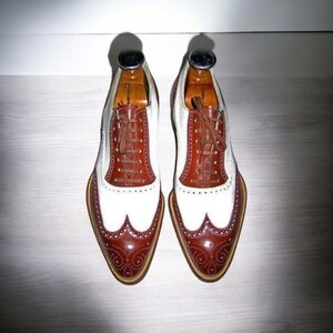 Puede incluir: Un par de zapatos de cuero marr&oacute;n y blanco con punta de ala y cordones. Los zapatos tienen una puntera blanca y una parte superior de cuero marr&oacute;n con un dise&ntilde;o de punta de ala. Los zapatos est&aacute;n sobre una superficie de madera.