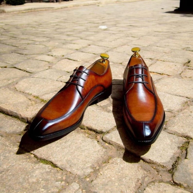 May include: A pair of brown leather lace-up shoes with a pointed toe and a contrasting darker brown leather panel on the toe. The shoes are on a brick surface.