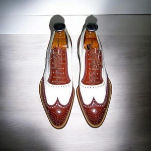 Puede incluir: Un par de zapatos de cuero marr&oacute;n y blanco con punta de ala y cordones. Los zapatos tienen una puntera blanca y una parte superior de cuero marr&oacute;n con un dise&ntilde;o de punta de ala. Los zapatos est&aacute;n sobre una superficie de madera.
