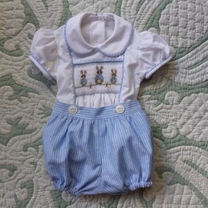 May include: A white and blue baby romper with a Peter Pan collar and short sleeves. The top has a smocked panel with embroidered bunnies and carrots. The bottom is blue and white striped with two white buttons.