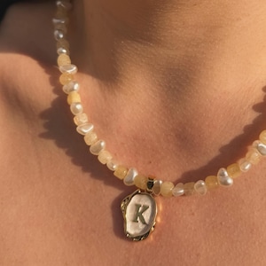 May include: A gold necklace with white pearl and yellow stone beads. The necklace has a gold pendant with a white background and the letter "K" in gold.