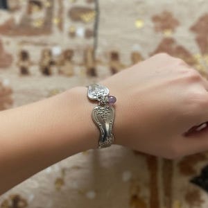 Peut inclure: Un bracelet en argent fabriqué à partir de couverts recyclés. Le bracelet présente un motif floral décoratif et une petite perle violette. Le bracelet est porté au poignet, mettant en valeur son style unique et artisanal. C'est un bijou unique.