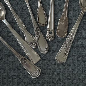 Op de afbeelding: Een assortiment antieke zilveren lepels met sierlijke bloemen- en krulwerkontwerpen. De lepels variëren in grootte en vorm, sommige met gedetailleerde patronen op de handvatten en bakken. Ze zijn gerangschikt op een getextureerd, donkergrijs oppervlak.