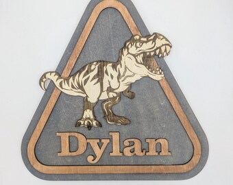 May include: A wooden triangle sign with a brown border and a gray background. A dinosaur silhouette is cut out of the wood and the name "Dylan" is written below the dinosaur.