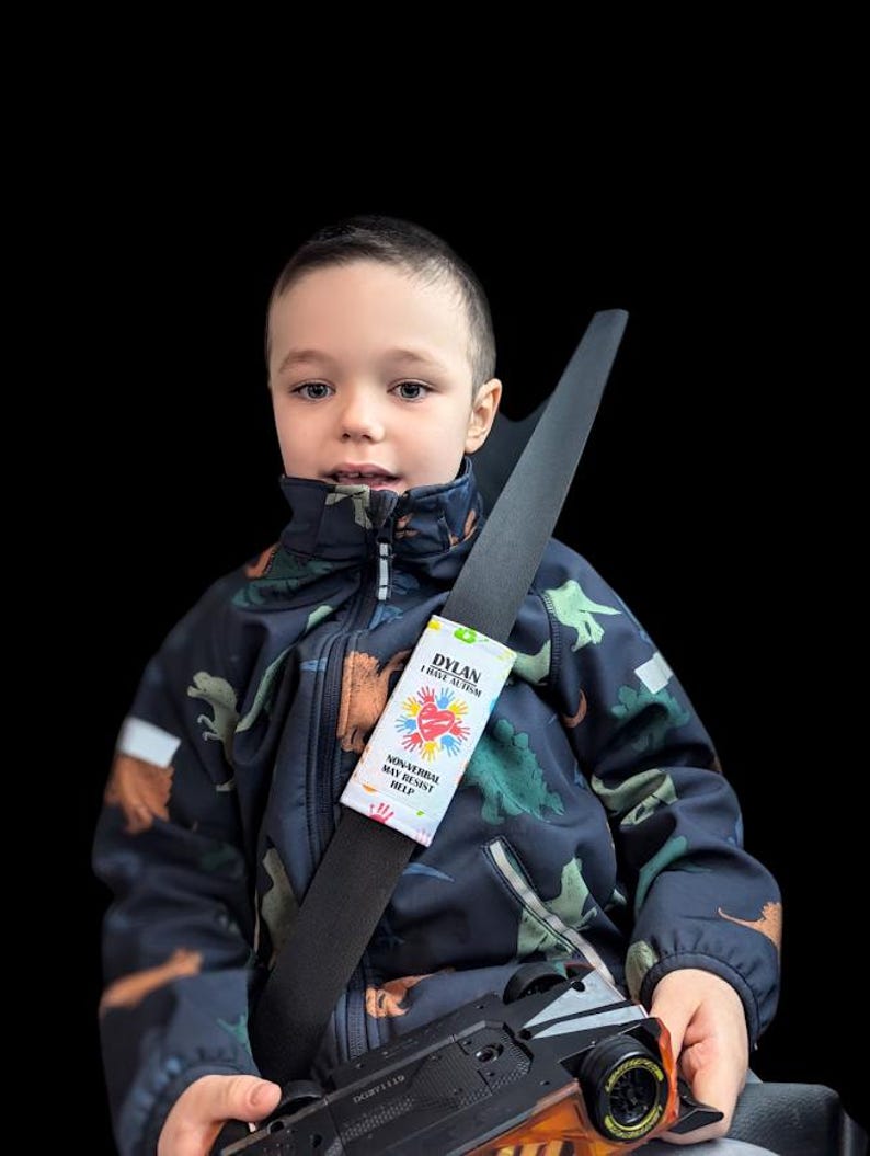 May include: A child wearing a navy blue dinosaur-patterned jacket with a seatbelt featuring a white label. The label reads "DYLAN I HAVE AUTISM" with a colorful heart and hand design, and the text "NON-VERBAL MAY RESIST HELP."