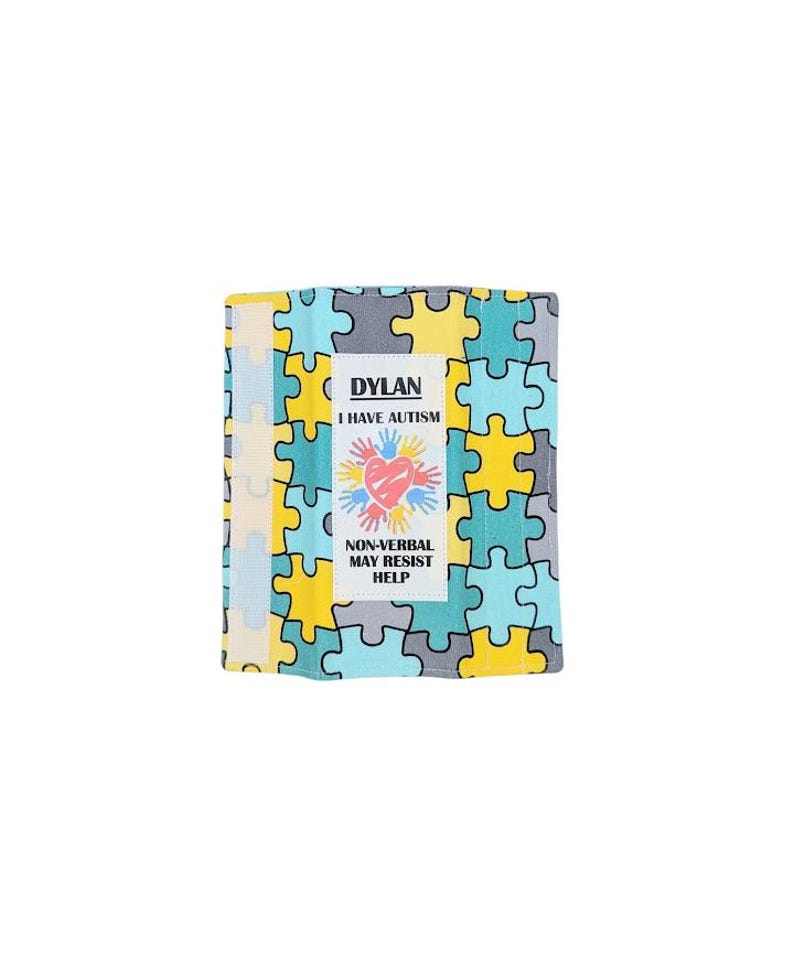 May include: A colorful, folded fabric item with a puzzle piece pattern in yellow, blue, gray, and cream. The item has a white label with the name "DYLAN", the text "I HAVE AUTISM", a heart with handprints, and the text "NON-VERBAL MAY RESIST HELP".