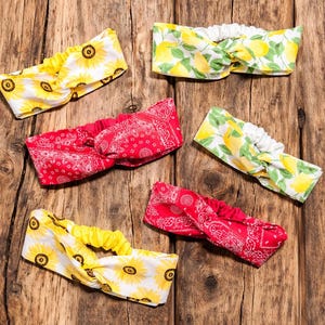 May include: Assortment of headbands with various patterns, including sunflowers, lemons, and red paisley. The headbands are made of fabric and have a twisted knot design. They are displayed on a rustic wooden surface.