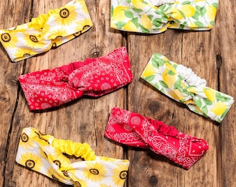 May include: Assortment of headbands with various patterns, including sunflowers, lemons, and red paisley. The headbands are made of fabric and have a twisted knot design. They are displayed on a rustic wooden surface.