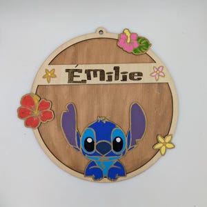 May include: A round wooden sign with a blue Stitch character from Lilo & Stitch. The sign is decorated with flowers and the name "Emilie" is written on the top.