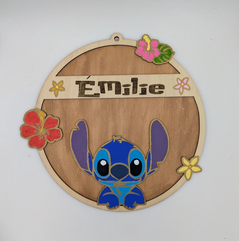 May include: A round wooden sign with a blue Stitch character from Lilo & Stitch. The sign is decorated with flowers and the name "Emilie" is written on the top.