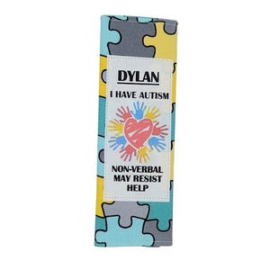 May include: A rectangular fabric item with a puzzle piece pattern in yellow, gray, and teal. A white label displays the name "DYLAN", the text "I HAVE AUTISM", and a graphic of a heart and hands. The text "NON-VERBAL MAY RESIST HELP" is also present.
