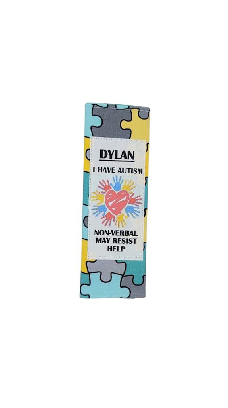 May include: A rectangular fabric item with a puzzle piece pattern in yellow, gray, and teal. A white label displays the name "DYLAN", the text "I HAVE AUTISM", and a graphic of a heart and hands. The text "NON-VERBAL MAY RESIST HELP" is also present.