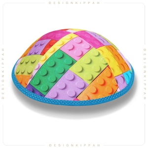 Può includere: Una kippa colorata con un motivo di blocchi Lego colorati. La kippa ha un bordo blu.